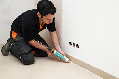 A man is kneeling and applying sealant to the base of a wall.