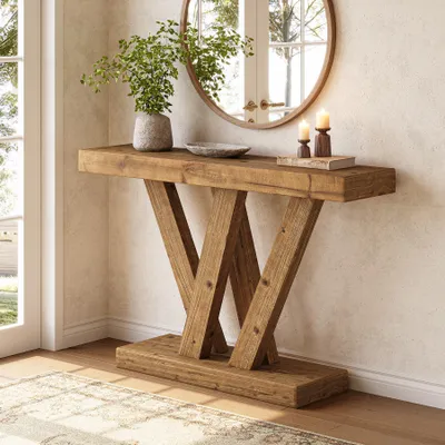 A rustic wooden console table with a V-shaped base is placed in a well-lit entryway or living room, next to a window and a round mirror.