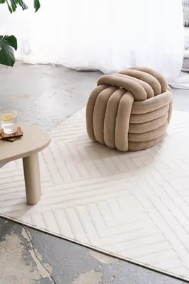 A beige knot pouf sits on a white geometric rug next to a small side table with a glass of water.