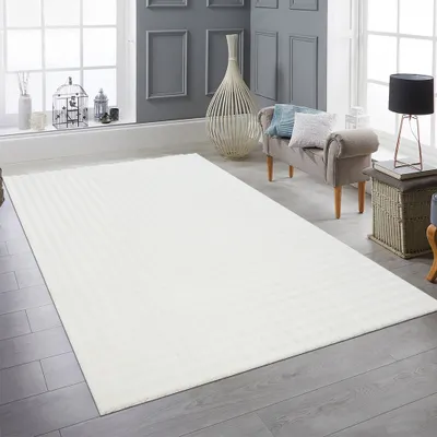 A cream-colored rug is laid out on a wooden floor in a room with modern decor. The rug is the main focus, with furniture and decorative items in the background.