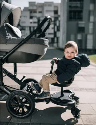 A baby sits on a stroller attachment while a stroller is beside them.