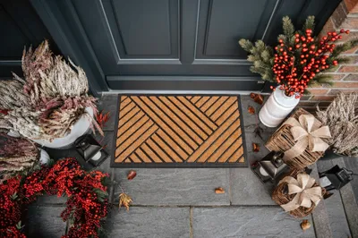 A doormat is placed in front of a dark gray door, decorated with festive items like a wreath, berries, and gifts.