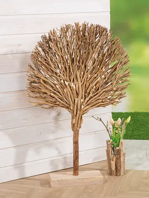 A decorative wooden tree sculpture stands on a light-colored base against a white wooden wall. To the right, a small vase made of driftwood holds some pink flowers and greenery.