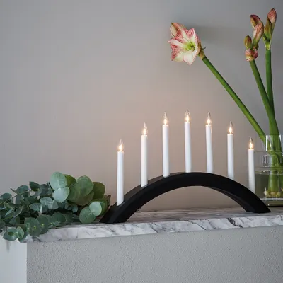 A black candle arch with seven white candles on a marble surface, with eucalyptus leaves and flowers in the background.