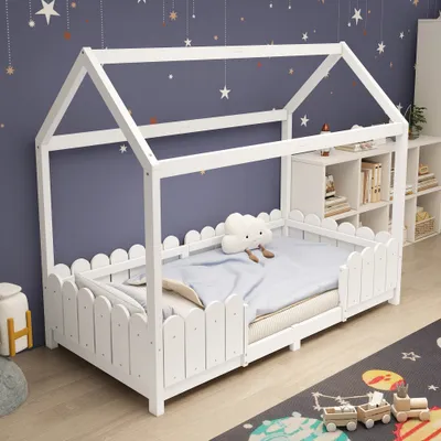 A white wooden house-shaped bed with a picket fence-like railing, set in a child's bedroom with star-themed wallpaper and a rug with planets and a toy train.