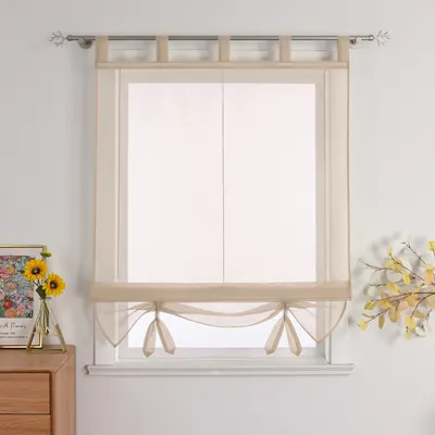 A sheer roman blind with tie-up details hangs in a window, with decorative items on a dresser to the left and a floral arrangement to the right.