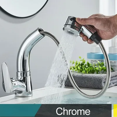 A hand is holding a pull-out faucet head, with water spraying into a sink. The faucet is chrome and has a modern design. There are also some toiletries and a plant in the background.