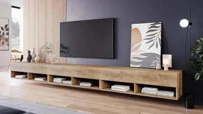 A modern living room with a TV mounted on the wall above a long, wooden media console. The console has several open compartments displaying books and decorative items. A piece of abstract art hangs on the wall next to the TV, and a floor lamp is positioned
