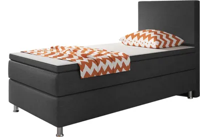 A single bed with a dark grey base and headboard, a white mattress, and a patterned throw blanket and pillow in orange and white.