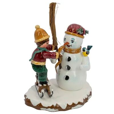 Ein Junge mit Schlittschuhen schmückt einen Schneemann mit einem Besen während ein kleiner Vogel auf dem Arm des Schneemanns sitzt und die Szene auf einer schneebedeckten Basis platziert ist