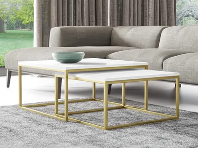 Two nesting coffee tables with white tops and gold metal frames are placed on a gray rug in front of a sectional sofa. A green bowl sits on the larger table.