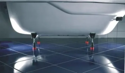 The underside of a white bathtub is shown, supported by four adjustable metal legs with red caps, resting on a dark blue tiled floor.