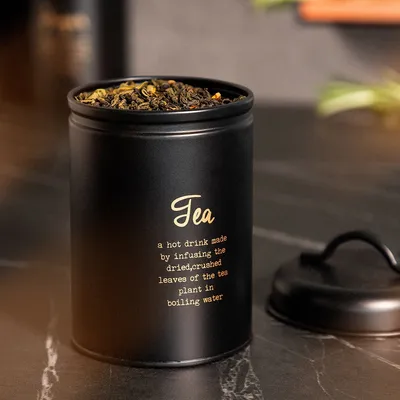 A black tea tin with gold lettering and dried tea leaves visible at the top, placed on a dark countertop.