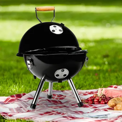 A black charcoal grill is set up on a picnic blanket in a grassy outdoor area, with some fruit and pastries nearby.