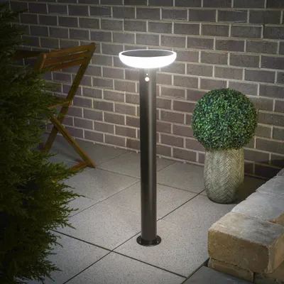 A solar-powered bollard light is shown on a patio at night, with a brick wall and some greenery in the background.