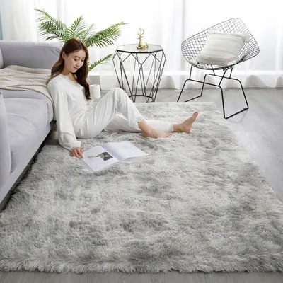 A woman is sitting on a fluffy gray rug in a living room, reading a book.