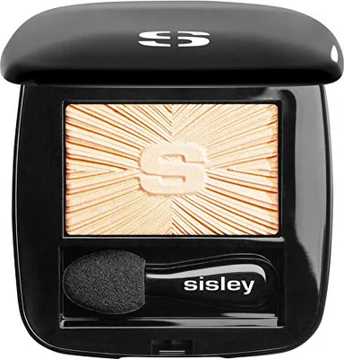 A black compact case containing a golden eyeshadow with a sunburst pattern and the brand's 'S' logo embossed in the center. An applicator with the brand name 'sisley' is also visible.