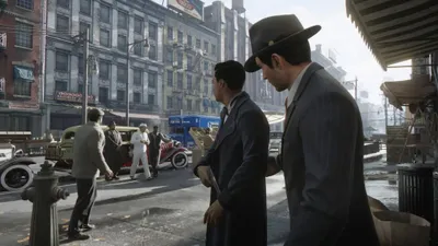 Two men in suits stand on a street corner in a city, with vintage cars and buildings in the background.