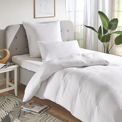 A neatly made bed with white bedding, pillows, and a duvet, set against a grey headboard and a window with sheer curtains and a plant.