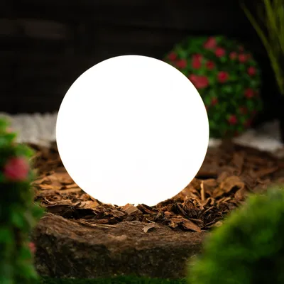 A glowing white sphere light sits on a bed of bark and rocks in a garden setting.