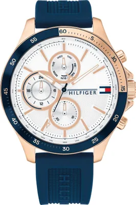 A close-up, front-facing studio shot of a Tommy Hilfiger chronograph watch with a white dial, rose gold case, and dark blue silicone strap.