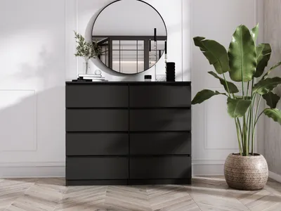 A black dresser with eight drawers is shown in a well-lit room with a round mirror above it and a large potted plant to the right.