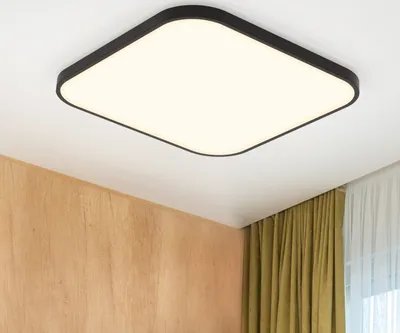 A square ceiling light with a black frame and a white illuminated panel is mounted on a white ceiling. The background shows a wall with wood paneling and green curtains.