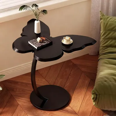 A black butterfly-shaped side table with a book, a small decorative house, a vase with a plant, and a cup of coffee on it, placed next to a green sofa.