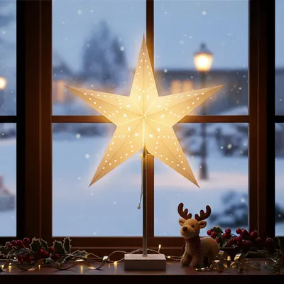 A lit paper star lantern sits on a windowsill in front of a snowy window, with a small reindeer figurine and festive decorations.