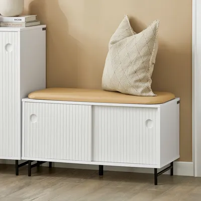 A white bench with a tan cushion and a decorative pillow sits in a room with a light beige wall and a wooden floor.
