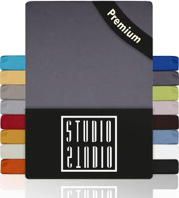 A stack of folded bed sheets in various colors, with a larger gray sheet prominently displayed in the foreground. A black band with the text 'Premium' is visible on the top right corner of the gray sheet. A black box with a white logo is at the bottom of t