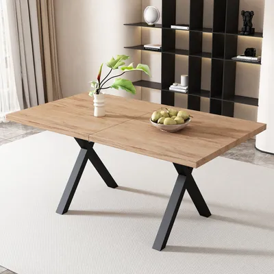 A modern dining table with a wooden top and black X-shaped legs, set on a light-colored rug. A vase with greenery and a bowl of fruit are on the table. A modern shelving unit is in the background.