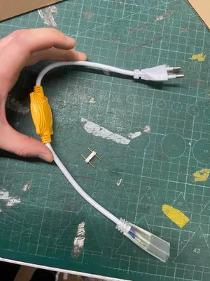A hand holding a white power adapter with a yellow connector, resting on a green cutting mat.