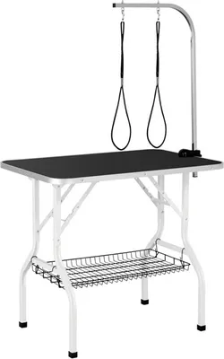A grooming table for pets, with a black tabletop, white foldable legs, and a silver grooming arm with two restraints. A wire basket is attached underneath the tabletop.