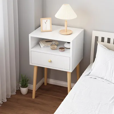 A white nightstand with a drawer and an open shelf, placed next to a bed. The nightstand has wooden legs and a small gold knob on the drawer. On the shelf, there are books, a perfume bottle, and a small vase. A lamp with a pleated shade sits on top of the