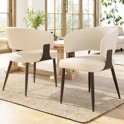 Two beige dining chairs with black legs are placed on a rug in front of a wooden dining table, with a vase of greenery on the table and a window in the background.