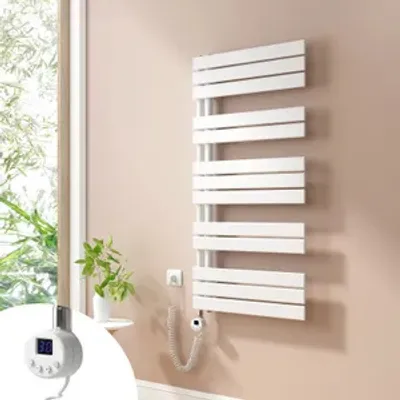 A white electric towel radiator mounted on a light beige wall next to a window with greenery visible outside. A small digital controller is attached to the radiator.