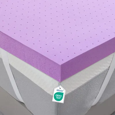 A close-up, angled view of a mattress topper with a purple foam layer on top, resting on a mattress. The topper has small perforations and elastic straps on the sides. A tag with branding and certification information is attached to the topper.