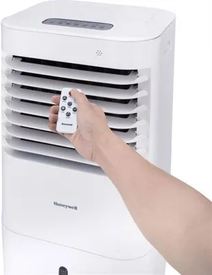 A hand holding a remote control in front of a Honeywell air cooler.
