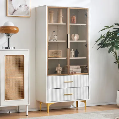 A white display cabinet with glass doors and two drawers, decorated with various items, stands in a room.