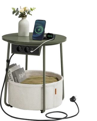 A round side table with integrated power outlets and USB ports, a phone and smartwatch charging, and a decorative vase with flowers on top. A storage basket with blankets is underneath.