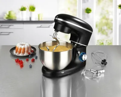 A black stand mixer with a stainless steel bowl filled with batter, placed on a kitchen counter next to a bundt cake and berries. The mixer comes with accessories like a whisk and dough hook.