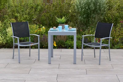 A small outdoor dining set with two chairs and a table on a patio, with greenery in the background.