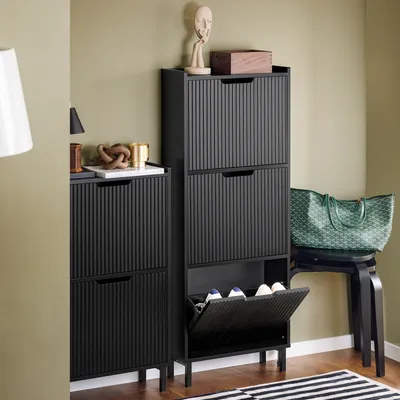 A black shoe cabinet with fluted doors is shown in a room with a light green wall and a wooden floor. A green tote bag is placed on a black stool next to the cabinet.