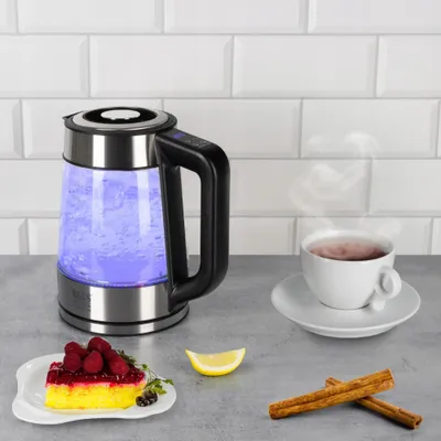 A glass electric kettle with blue light, a cup of tea, a slice of cake, lemon, and cinnamon sticks on a countertop.