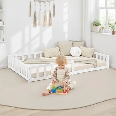A child plays with a stacking toy on the floor next to a white toddler bed with a guardrail.