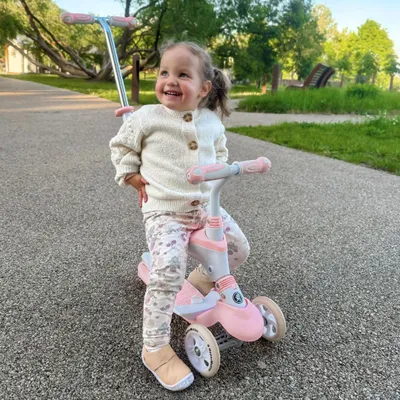 A young child is happily sitting on a pink and white balance scooter in a park.