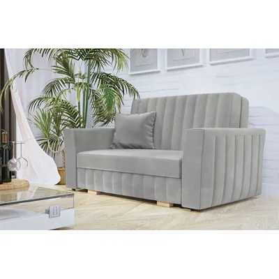 A light gray sofa with a single throw pillow is shown in a well-lit room with a large potted plant and sheer curtains in the background.