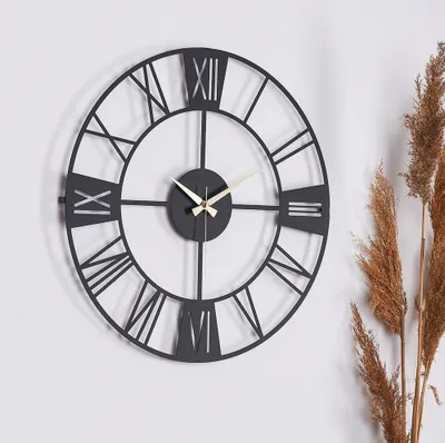 A black metal wall clock with Roman numerals is displayed on a white wall, next to dried grass.