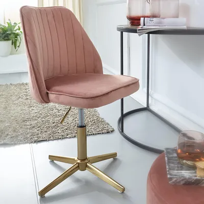 A pink velvet office chair with a gold base is shown next to a desk with decorative items.